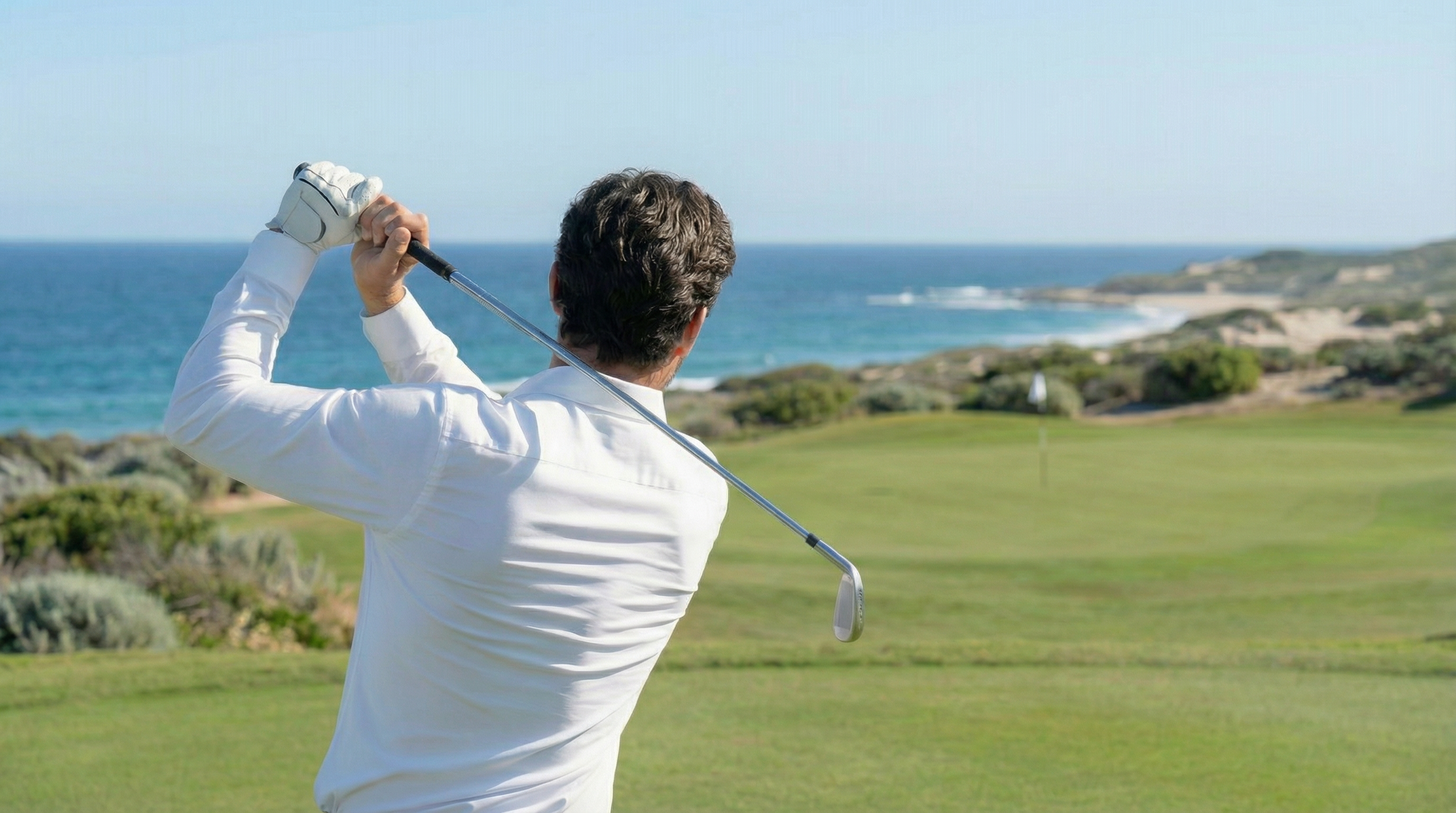 Homme en chemise blanche stretch réalisant un swing sur un parcours de golf en bord de mer.