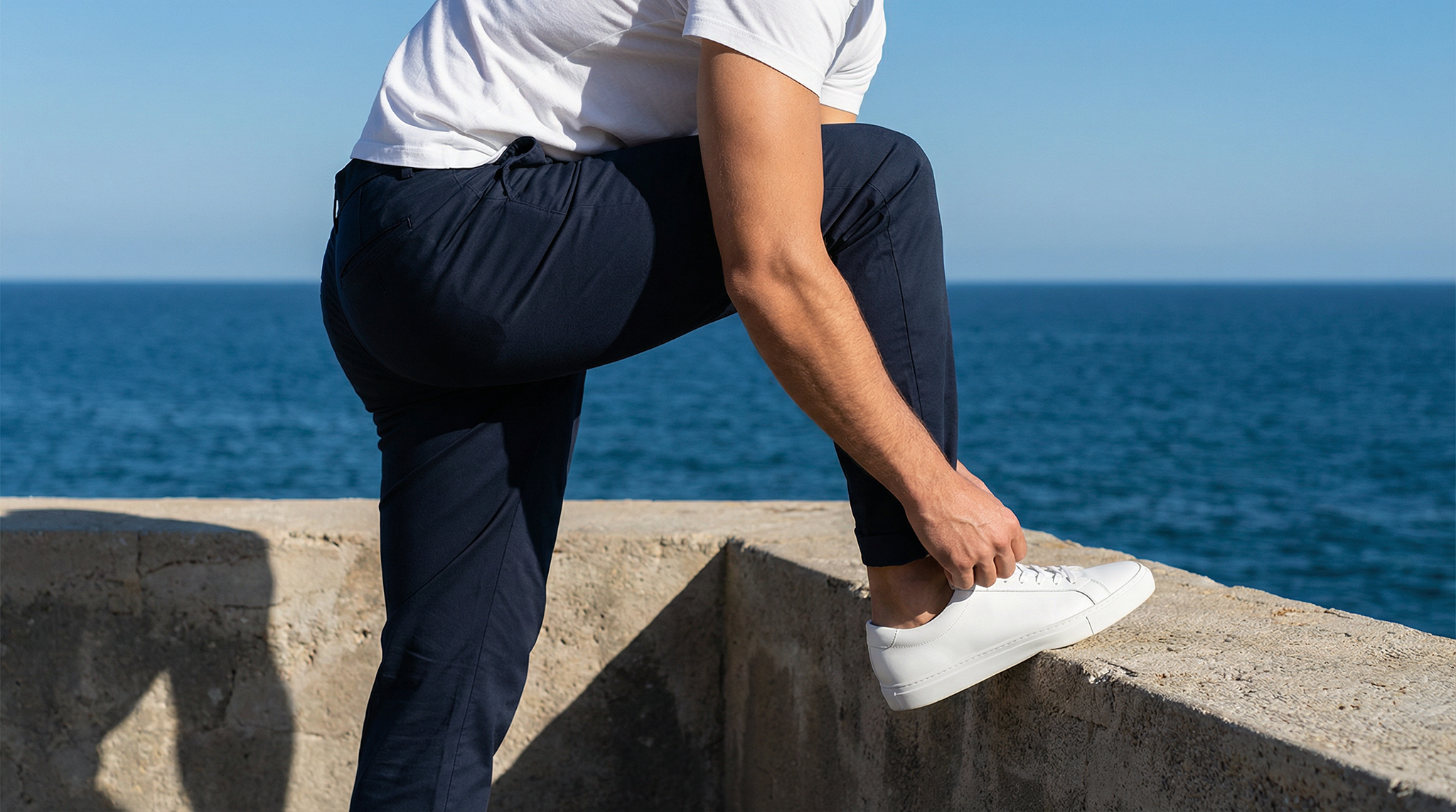 Homme portant un chino bleu marine et des sneakers blanches face à la mer.