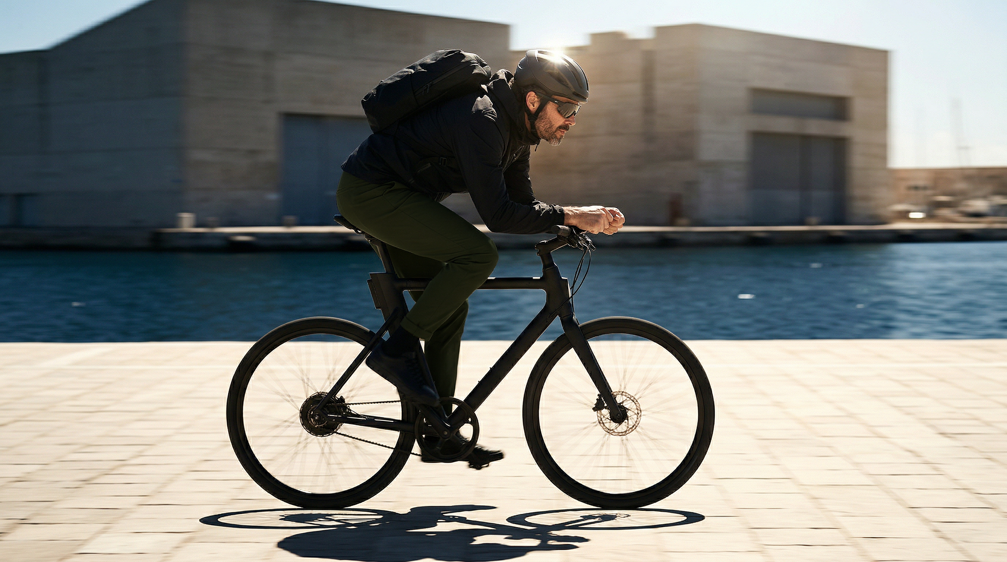 Cycliste portant un pantalon chino vert foncé et un sac à dos le long d’un quai maritime.