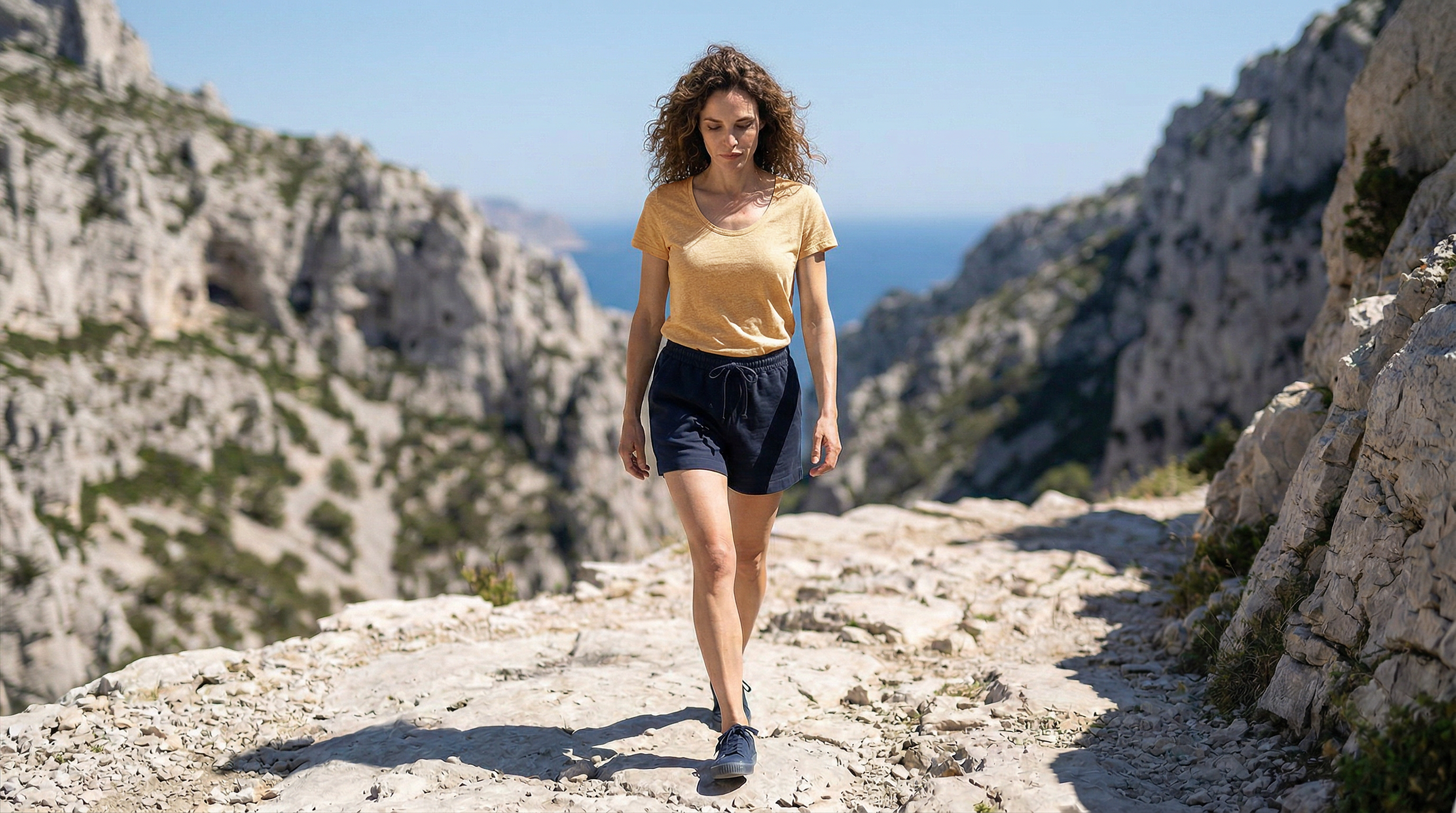 Silhouette féminine en t-shirt ocre col rond évoluant sur un chemin minéral entre falaises.