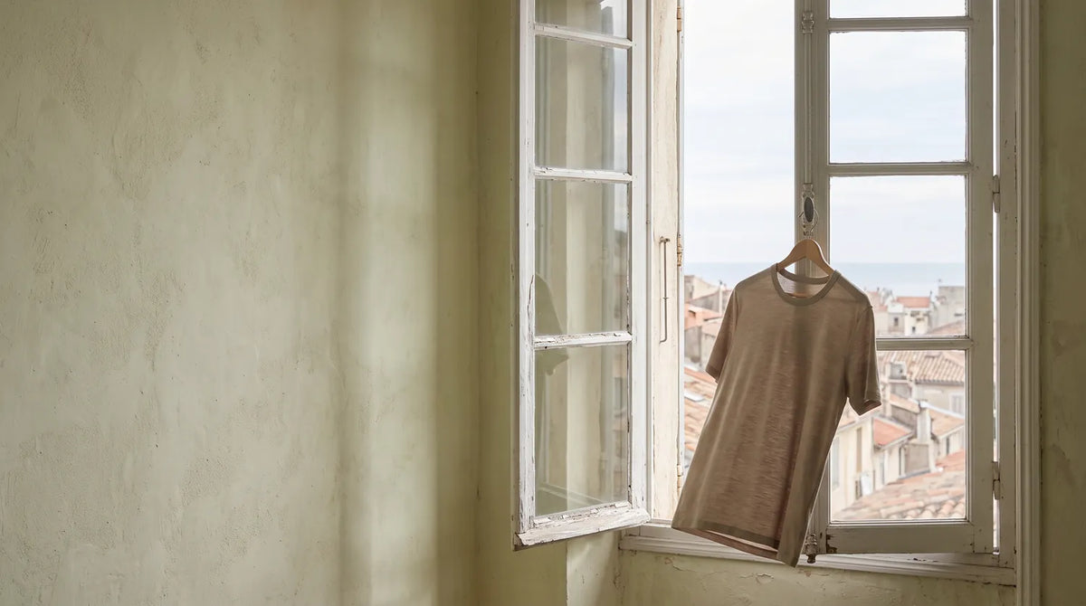 A blue Performance Merino T-shirt on a hanger, hooked to the latch of a wide-open window, in the evening