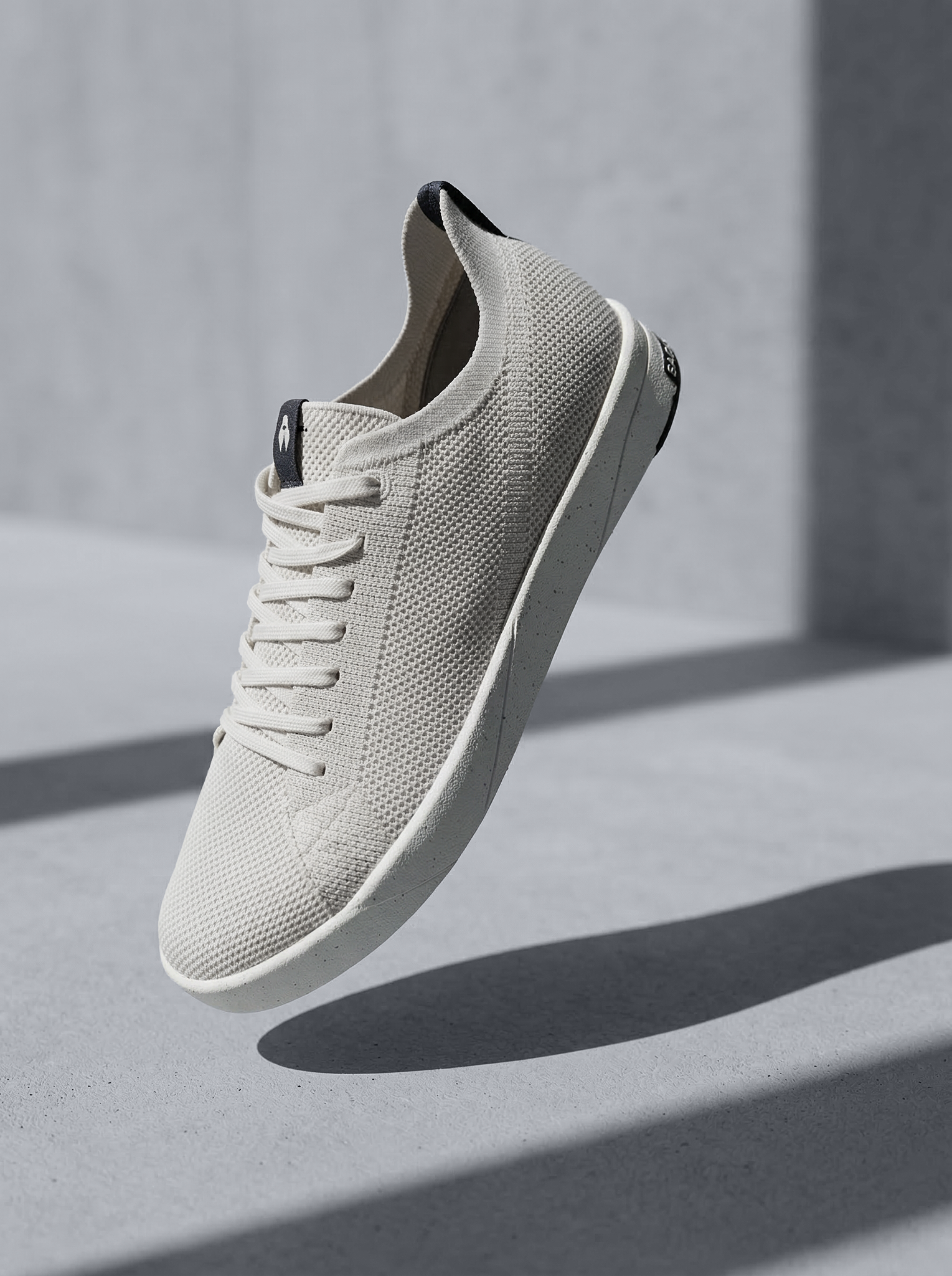 Sneaker beige en maille respirante suspendue dans l’air, ombre graphique sur fond minéral.