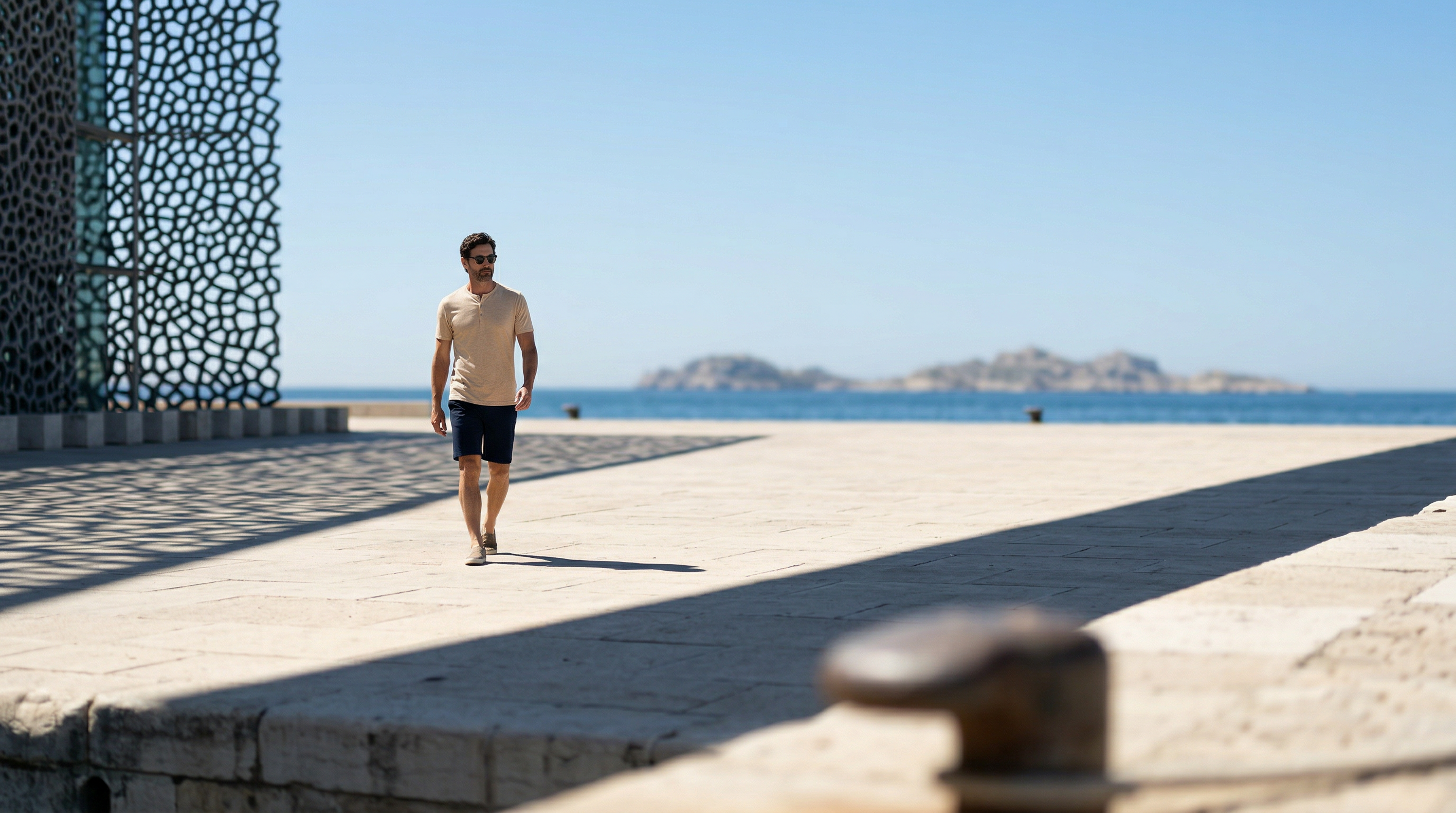 Silhouette masculine élégante en t-shirt beige avançant sur un quai ensoleillé au bord de l’eau.