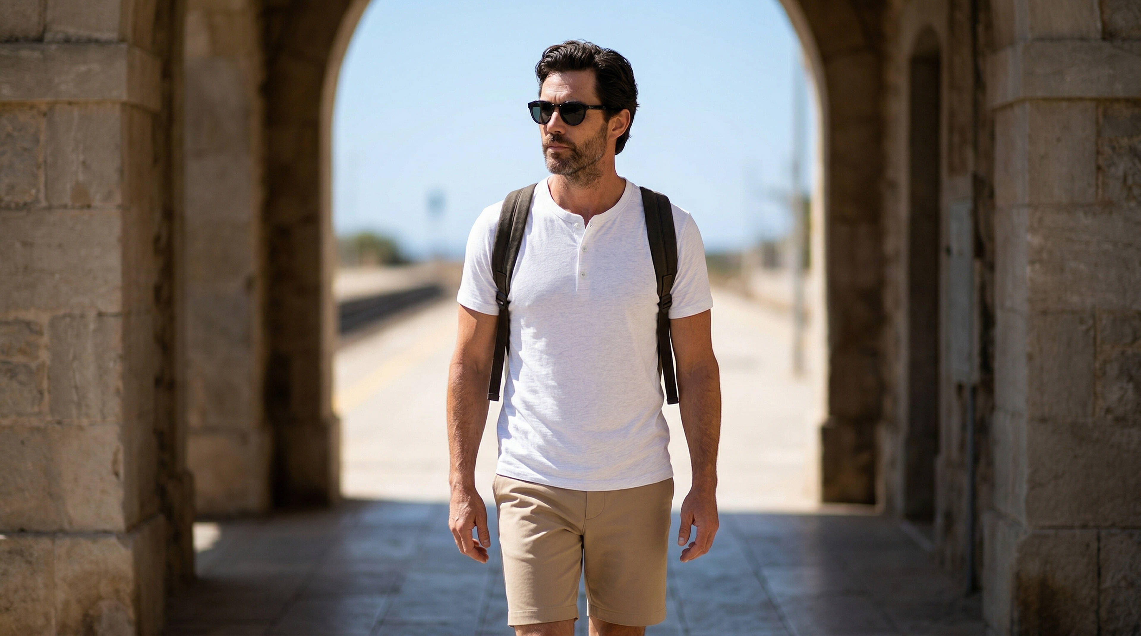 Silhouette masculine élégante en Henley blanc traversant une arche méditerranéenne ensoleillée.
