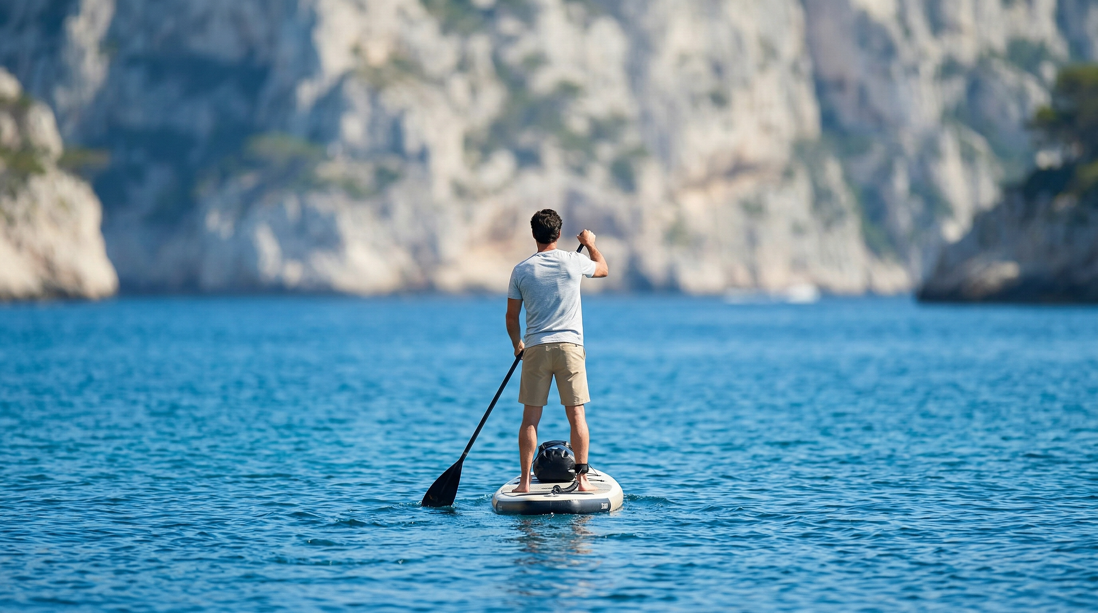 Silhouette masculine en t-shirt gris pagayant sur une mer turquoise au pied de falaises calcaires.
