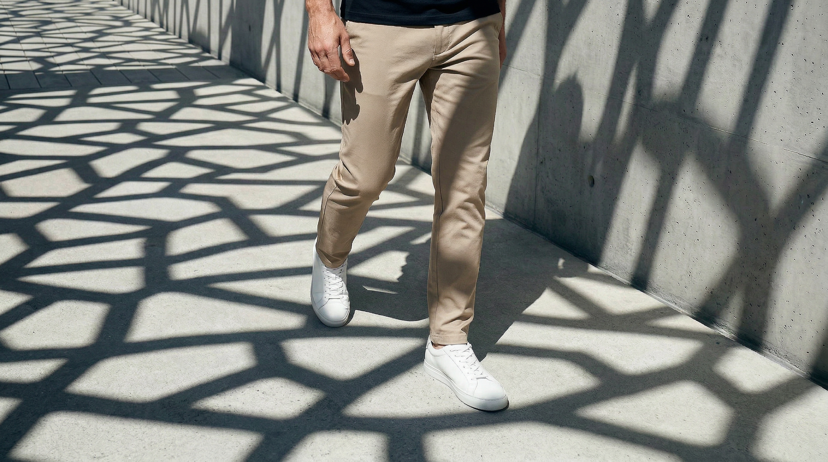 Détail d’un pantalon chino beige porté avec des sneakers blanches dans un environnement architectural moderne.