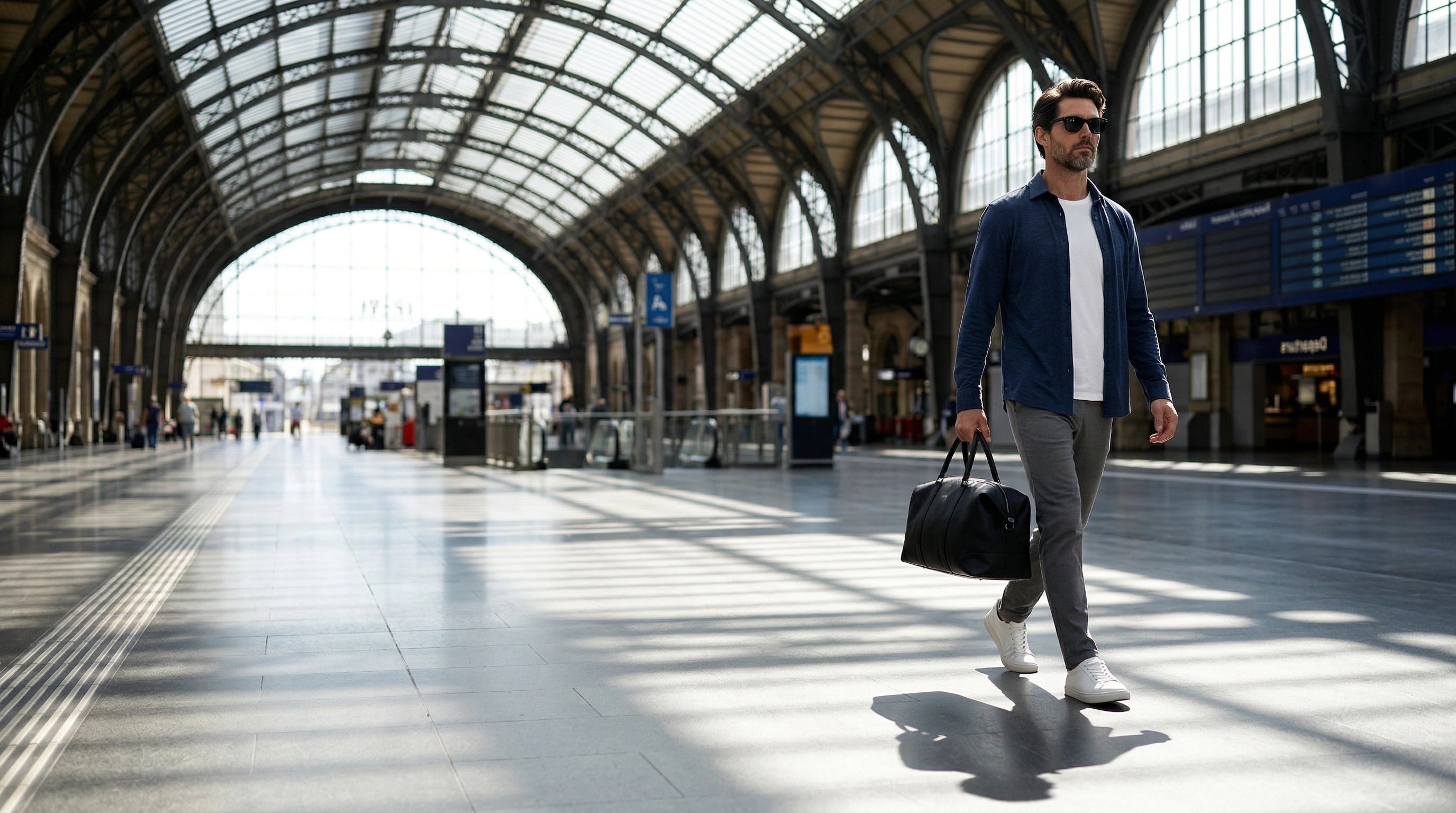 Homme portant des lunettes de soleil noires marchant dans une gare avec un sac de voyage à la main.