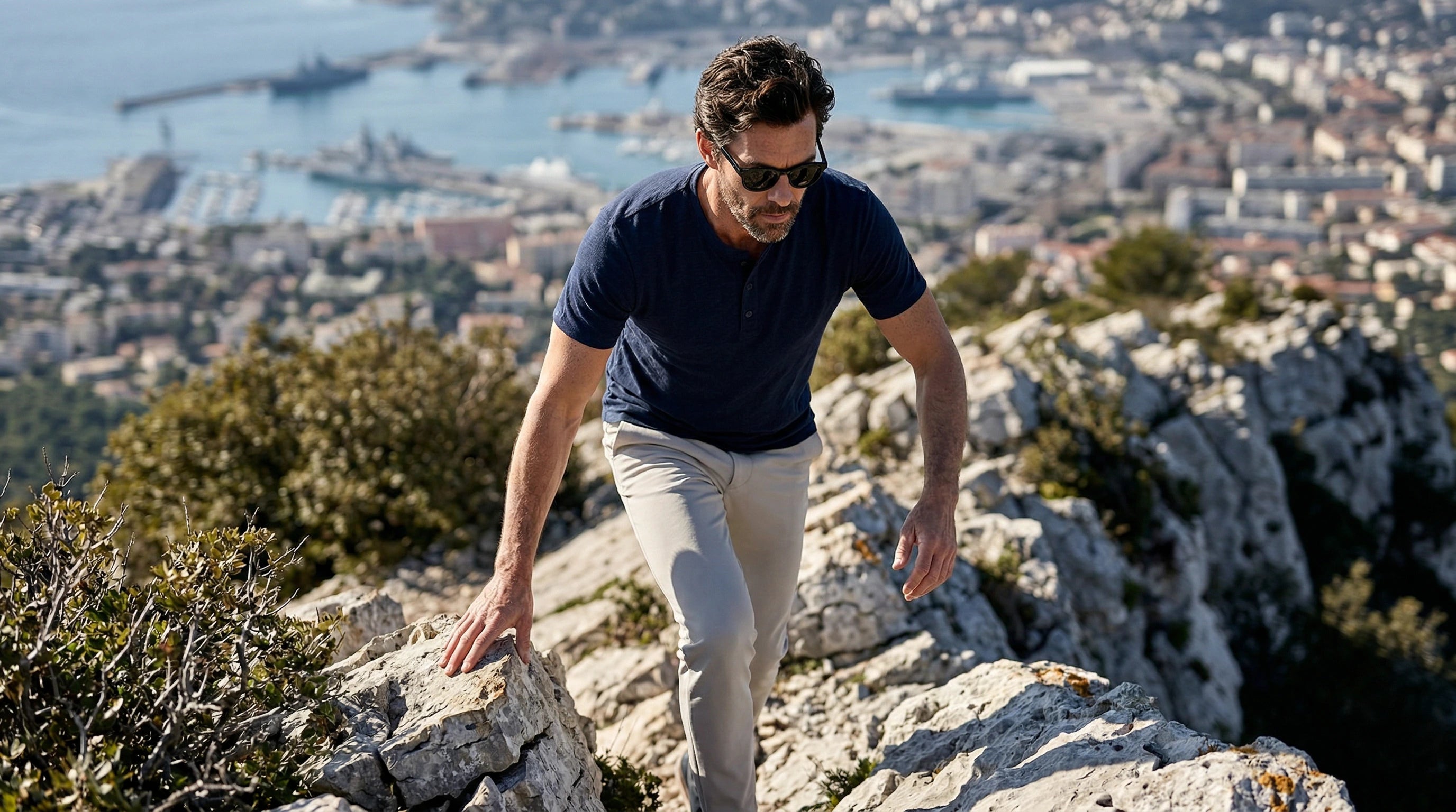 Homme portant des lunettes de soleil noires lors d’une randonnée avec vue panoramique sur la mer et la ville.
