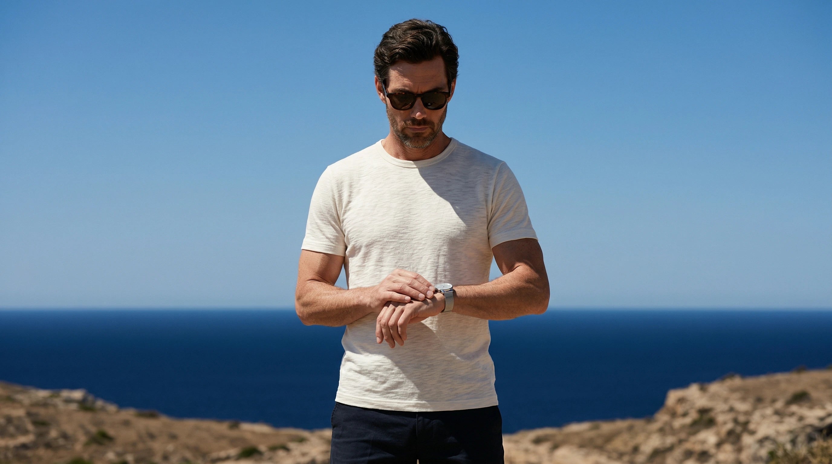 Homme portant des lunettes de soleil premium en bord de mer, consultant sa montre face à l’horizon méditerranéen.