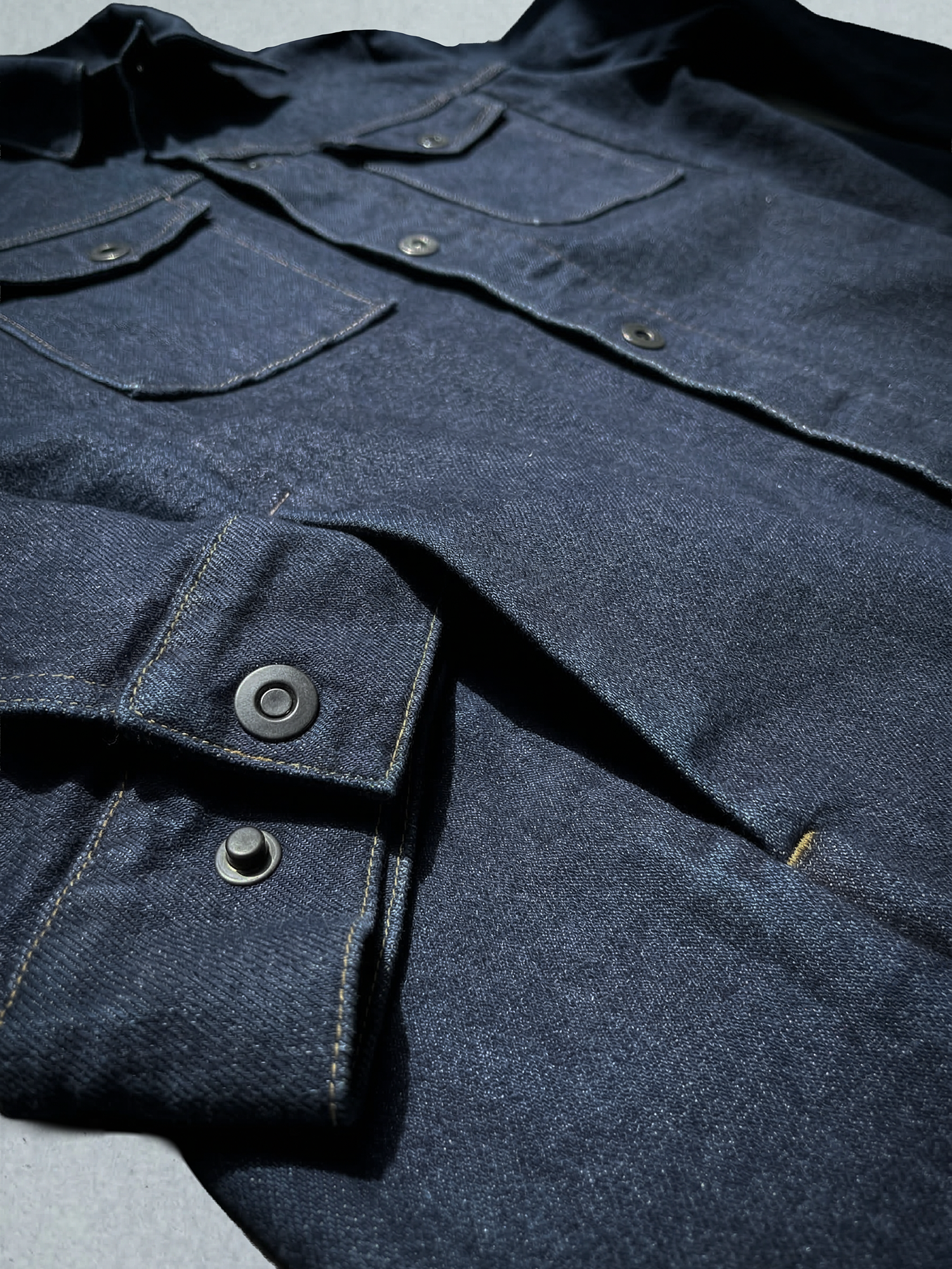Détail d’une veste en jean bleu foncé avec coutures contrastées et boutons métalliques.