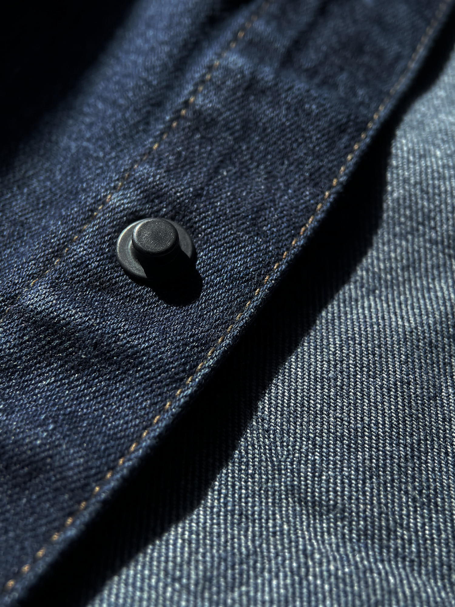 Gros plan sur un bouton pression métallique noir avec coutures contrastées sur veste en jean.